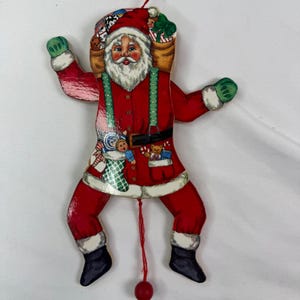 May include: A wooden Santa Claus jumping jack ornament featuring a red suit with white trim, green braces, and black boots. Santa carries a sack of toys on his back and has a small doll and teddy bear graphic on his coat. A red string hangs below.