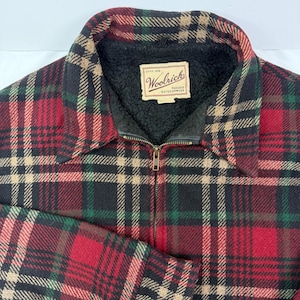 May include: A zip-up jacket featuring a red, black, green, and tan plaid pattern. The interior is lined with black sherpa fleece. A rectangular label at the neckline reads Woolrich Since 1830 Rugged Outdoorwear in black text on a cream background.
