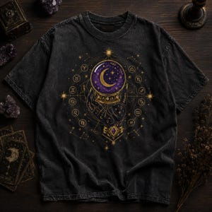 May include: A charcoal black short-sleeve t-shirt featuring a central graphic of a hand holding a purple crystal ball with a crescent moon and stars. The design is surrounded by a gold circular zodiac wheel, celestial patterns, and stars.