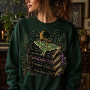 Pode incluir: Uma camisola verde-escura com uma impressão gráfica de uma pilha de quatro livros de estilo vintage intitulados Luna Mystica, Nocturna Naturae, Herbarium e Astraea Vol. 1, encimada por uma grande traça-luna verde, lua crescente e cristais roxos.