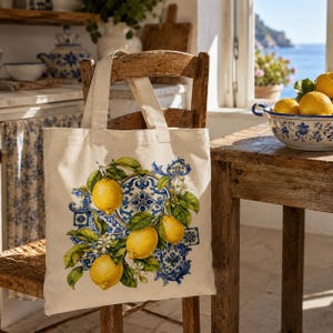 May include: A cream-colored canvas tote bag featuring a printed design of three bright yellow lemons with green leaves and white blossoms, layered over a decorative blue and white tile pattern. The bag has two sturdy fabric handles.