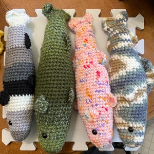 May include: Four handmade crochet fish toys arranged side by side. The first is gray, white, and black striped. The second is solid olive green. The third is light pink with multicolored speckles. The fourth features a blue, tan, and white camouflage pattern.