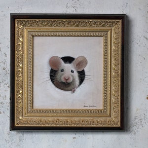 Puede incluir: Una pintura enmarcada de un rat&oacute;n blanco asom&aacute;ndose por un agujero. El rat&oacute;n tiene orejas rosas y una nariz negra. El marco es dorado con un borde marr&oacute;n oscuro.