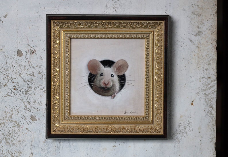Puede incluir: Una pintura enmarcada de un rat&oacute;n blanco asom&aacute;ndose por un agujero. El rat&oacute;n tiene orejas rosas y una nariz negra. El marco es dorado con un borde marr&oacute;n oscuro.