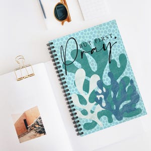 May include: A spiral-bound notebook featuring a light blue background with a dark blue polka dot pattern and abstract teal and white coral-like shapes. The text "BUT FIRST PRAY" is printed in black script and sans-serif font on the front cover.