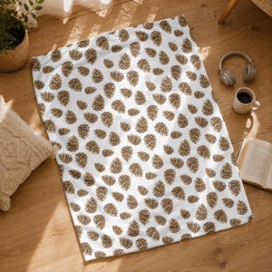 Puede incluir: Una manta rectangular blanca con un patrón repetitivo de piñas marrones distribuidas por toda la superficie. La tela tiene una textura suave y muestra un estampado consistente de formas de piñas naturales en diversas orientaciones.