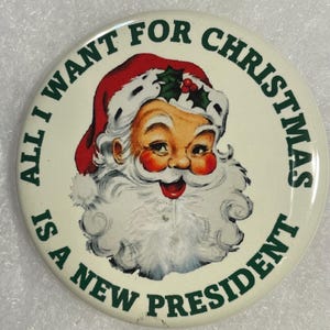 May include: A round button with a Santa Claus graphic. Santa is wearing a red hat with white trim and a white beard. The text around the image reads "ALL I WANT FOR CHRISTMAS IS A NEW PRESIDENT" in green.
