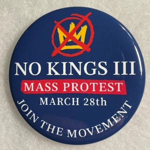 May include: A round, navy blue button with the text "NO KINGS III MASS PROTEST MARCH 28th JOIN THE MOVEMENT". The button features a red crossed-out crown design and white text.