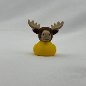 Peut inclure: Une petite figurine représentant la tête d'un élan brun avec des bois beige clair fixée sur un corps de canard en caoutchouc jaune vif. L'élan a des yeux sombres et un museau brun clair, tandis que le corps du canard est lisse, arrondi et jaune.
