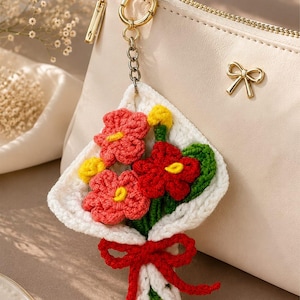 May include: A crochet flower bouquet keyring featuring red and coral blossoms with yellow centres, green stems, and a white wrap tied with a red yarn bow. The keyring is attached to a gold-toned metal ring and clip, hanging from a beige zippered pouch.