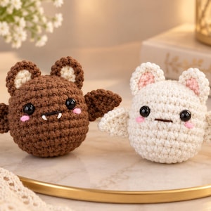 May include: Two small, round, crocheted bat plushies. One is dark brown with cream-colored inner ears and white fangs, while the other is cream-colored with light pink inner ears. Both have black plastic eyes and pink stitched cheeks.