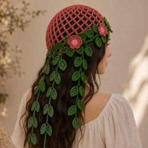 May include: A crochet cap in a dusty rose color featuring a lattice pattern. The base of the cap is adorned with a border of green crocheted leaves and small pink flowers, with several long, trailing green vine strands hanging down the back.