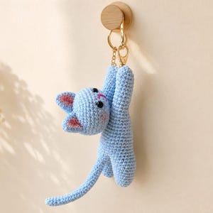 May include: A light blue crocheted cat keychain featuring a small stuffed feline figure with black bead eyes, pink stitched nose and mouth, and pink inner ears. The cat is attached to a gold-toned metal lobster clasp and ring for hanging.