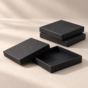 May include: Three square black cardboard boxes with matte finishes. One box is open, revealing a hollow interior, while the other two are closed and stacked. The boxes feature clean, sharp edges and a simple, uniform design for storage or gift packaging.