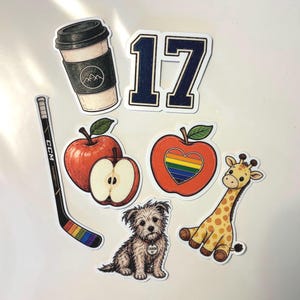 May include: A collection of stickers featuring a coffee cup, the number 17 in navy blue, a hockey stick with rainbow tape, a whole red apple, a sliced apple, an apple with a rainbow heart, a small brown dog, and a yellow giraffe with brown spots.