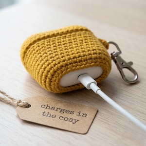 May include: A mustard yellow crocheted protective case for wireless earbuds, featuring a silver metal lobster clasp attached to the side. A white charging cable is plugged into the bottom of the case, and a brown paper tag reads charges in the cosy.