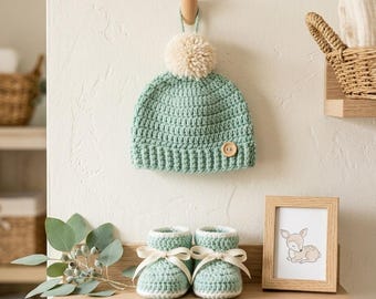 May include: A light green crocheted beanie with a cream-coloured pom-pom and a single wooden button, paired with matching light green crocheted booties featuring cream-coloured ribbon bows. The set is displayed on a light-coloured wooden shelf.
