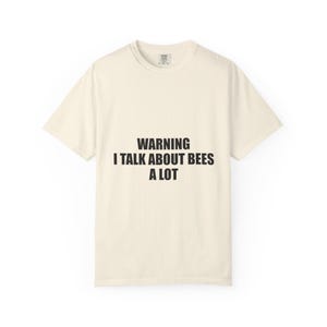 Può includere: Una t-shirt a maniche corte color crema con il testo nero "WARNING I TALK ABOUT BEES A LOT" stampato al centro del petto. Il capo presenta uno scollo rotondo e una vestibilità standard, con un'etichetta del marchio visibile all'interno del colletto.