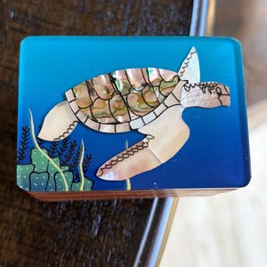May include: A rectangular wooden box with a blue top featuring a sea turtle design. The turtle's shell is made of iridescent mother-of-pearl, and the box has a marine theme with seaweed accents.