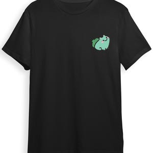 Könnte beinhalten: Ein schwarzes Kurzarm-T-Shirt mit einer kleinen Grafik auf der linken Brustseite. Die Grafik zeigt ein stilisiertes, hellgrünes Wesen mit einer dunkelgrünen, zwiebelförmigen Struktur auf dem Rücken, einem lächelnden Gesicht und rosa Wangen.