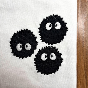 May include: A white fabric surface featuring three embroidered black circular shapes with jagged edges. Each shape contains two large, round white eyes with black pupils, creating a cluster of three distinct, fuzzy-looking characters.