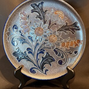 Peut inclure: Une assiette ronde en céramique avec une finition mouchetée bleu foncé et gris et un motif floral en relief. Le centre présente des motifs botaniques complexes en relief, incluant des fleurs et des feuilles stylisées dans des tons de bleu, brun et gris.