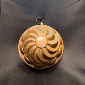 Op de afbeelding: Een ronde koperen vorm met een wervelend, spiraalvormig patroon dat uitstraalt vanuit een glad middelpunt. Het metalen oppervlak vertoont een warme, verouderde patina met zichtbare kleurvariaties. Een kleine metalen ring is aan de bovenkant bevestigd.