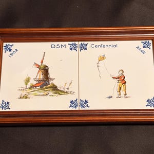 May include: A rectangular wooden frame containing two white ceramic tiles. The left tile features a windmill and the text 1902. The right tile shows a person flying a kite and the text 2002. The text DSM Centennial is centered above the tiles.