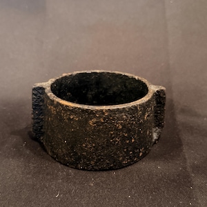 May include: A circular, dark brown, cast iron ring with a rough, textured surface. The object features two small, protruding rectangular tabs on opposite sides of the exterior wall, creating a simple, industrial, and heavy-duty appearance.