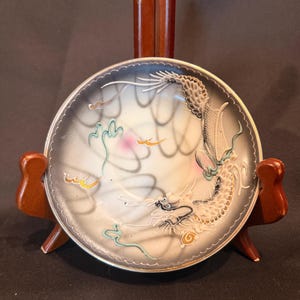 May include: A round ceramic dish featuring a raised, textured dragon motif in white and gold against a gradient gray background. The dragon curves along the edge, accented with small touches of pink, green, and gold. The dish has a scalloped rim.