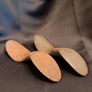 May include: Two cast iron cobbler shoe lasts with a weathered, oxidized surface. These tools feature a curved, elongated shape with a rounded heel and a tapered toe section, displaying a rough, textured patina consistent with aged metal hardware.
