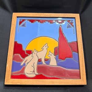 May include: A square decorative tile framed in light wood featuring a colourful enamel-style illustration of three coyotes howling at a large yellow sun. The scene includes red rock formations, purple ground, and a light blue sky with geometric accents.