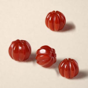 May include: Four small, round, deep red beads with a carved, vertical ribbed texture. Each bead features a central hole and a smooth, glossy finish, arranged in a scattered pattern on a light, neutral-colored surface.