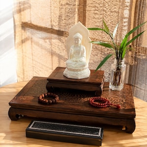 Puede incluir: Una estatua de Buda blanca sobre un pedestal de madera oscura colocado en una base rectangular mayor. Junto a ella hay un jarrón de cristal con hojas verdes, una pulsera de cuentas marrones, un mala de cuentas rojas y una caja de incienso negra.