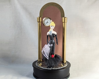 May include: A black and gold musical figurine of a Barbie doll singing on a stage with a microphone. The doll is wearing a black sequined dress and a white feather boa. The stage has a clock with the words "Solo in the Spotlight" above it.