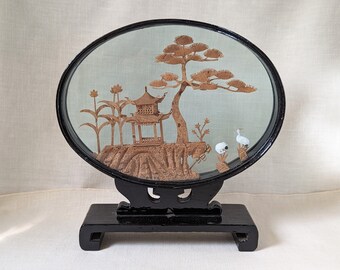 May include: A black framed oval glass display with a cork sculpture of a pagoda, a pine tree, and two birds. The display is on a black stand with a rectangular base.