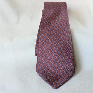 May include: A maroon tie with a repeating geometric pattern of interlocking shapes in shades of blue and purple. The tie is made of a smooth fabric and is displayed on a white surface.