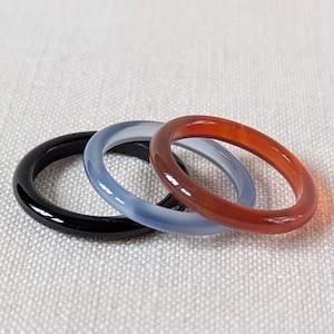 May include: Three gemstone rings in black, blue, and red-orange. The rings are smooth and polished, with a simple band design. The rings are displayed on a neutral, textured surface. The rings are likely for fashion or jewelry.