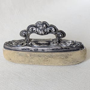 May include: Antique silver-toned metal sewing iron with a decorative handle and a rectangular base. The base is a light tan color. The handle features ornate scrollwork and floral designs. A vintage sewing tool.