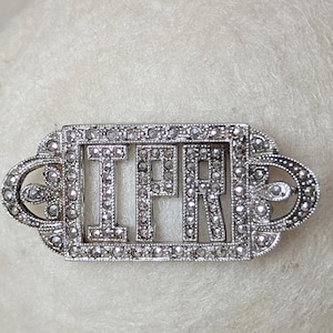 May include: A silver-toned vintage brooch with the letters "IFR" set within a rectangular frame. The frame and letters are encrusted with small, sparkling stones. The brooch has decorative, curved end pieces.