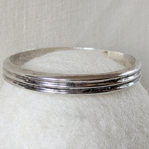May include: A silver-toned bangle bracelet with three parallel, textured bands. The bracelet is circular and appears to be made of metal. The jewelry is displayed on a white, textured surface.