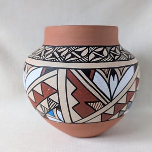 May include: A terracotta-colored ceramic pot with a rounded body and a short neck. The pot is adorned with a geometric pattern in black, white, and brown, featuring triangles, lines, and floral-like shapes. The design is symmetrical and covers the majority of the pot's surface.