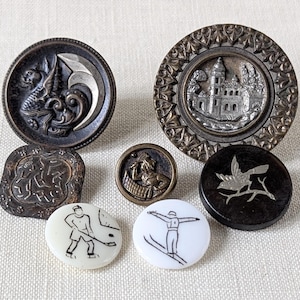 May include: A collection of antique buttons with various designs. Includes a dragon, a castle, a bird on a branch, a figure in a boat, and two buttons depicting hockey and skiing. The buttons are in shades of brown, silver, and white.