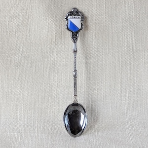 May include: A silver souvenir spoon with a blue and white enamel shield on the handle. The shield has the word "Zurich" written on it.