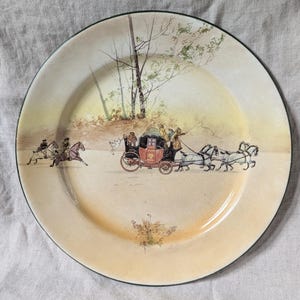 May include: A vintage ceramic plate with a cream-colored background and a green rim. The plate features a hand-painted scene of a horse-drawn carriage with passengers traveling through a wooded area.