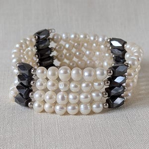 May include: A multi-strand bracelet featuring lustrous white pearls and geometric black hematite accents. The bracelet is composed of multiple rows of pearls, connected by the dark, faceted hematite stones. The design is elegant and suitable for various occasions.