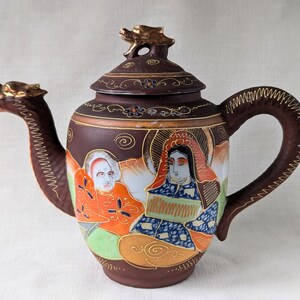 May include: A brown ceramic teapot with a dragon spout and handle. The teapot is decorated with gold accents and a painted scene of two figures in traditional Japanese clothing.