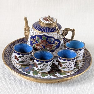 May include: A miniature tea set with a teapot and four cups, all resting on a round tray. The set features a white base with blue, red, and gold floral designs. The cups and teapot have blue interiors.