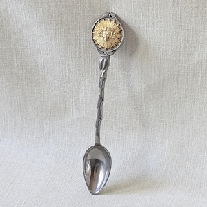 May include: A decorative spoon with a silver-colored bowl and a twisted stem. The spoon's handle features a gold-colored sunburst design with a face in the center. The spoon is displayed against a neutral background.