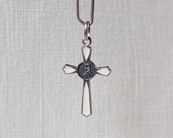 May include: A silver-toned cross pendant with a white enamel border and a dark blue center. The cross is suspended from a delicate silver chain. The center of the cross features a symbol. The background is a textured, off-white fabric.